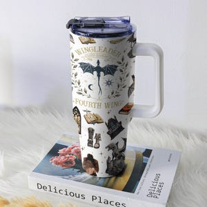 May include: White insulated tumbler with a handle, featuring a fantasy-themed design. The design includes a dragon, books, and the text "WINGLEADER, FOURTH WING". The tumbler is sitting on a book titled "Delicious Places".