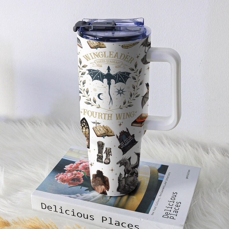 May include: White insulated tumbler with a handle, featuring a fantasy-themed design. The design includes a dragon, books, and the text "WINGLEADER, FOURTH WING". The tumbler is sitting on a book titled "Delicious Places".