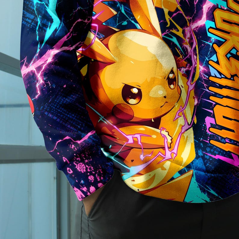 May include: A long-sleeve shirt featuring a vibrant graphic print of a yellow Pikachu character surrounded by bright pink and blue lightning bolts against a dark blue background. The design includes stylized orange text on the right side.