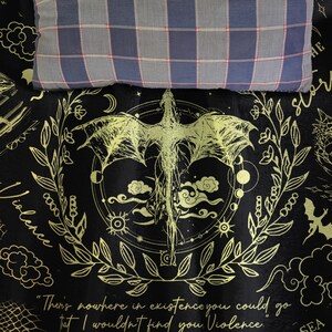 May include: Black and gold dragon-themed blanket with a quote about violence. The blanket features a dragon illustration, celestial elements, and a decorative border. A plaid pillow rests on top.