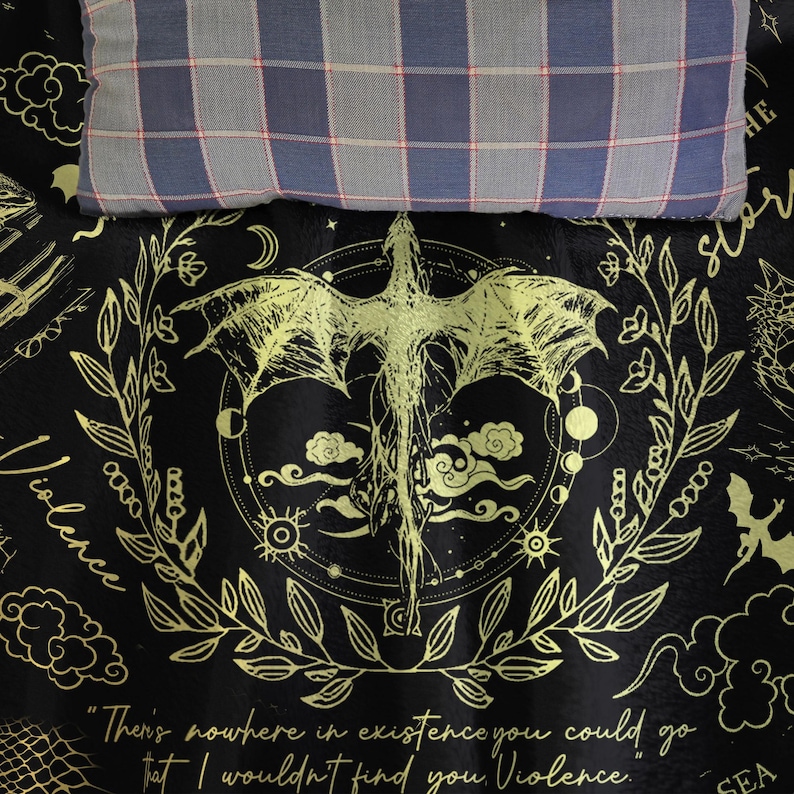 May include: Black and gold dragon-themed blanket with a quote about violence. The blanket features a dragon illustration, celestial elements, and a decorative border. A plaid pillow rests on top.