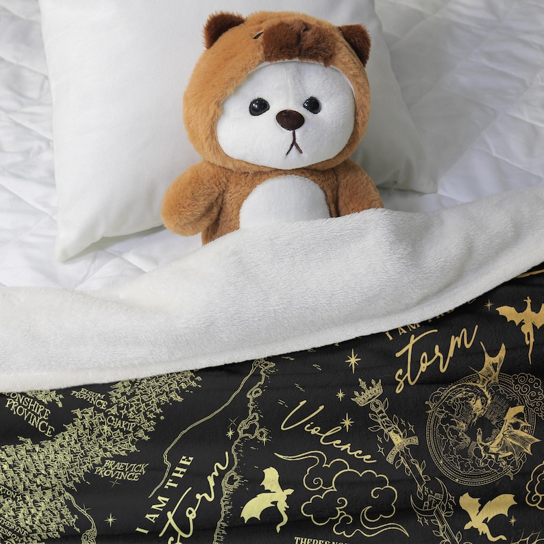 May include: A brown teddy bear wearing a bear costume is nestled under a white blanket on a bed. The blanket has a black background with gold illustrations and the words "I AM THE storm" and "Violence".