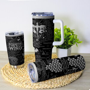 May include: Three black insulated tumblers with white graphic designs and text. One tumbler reads "FOURTH WING" with dragon illustrations. Another features a map design with the text "POROMIEL". The third has a scale pattern.