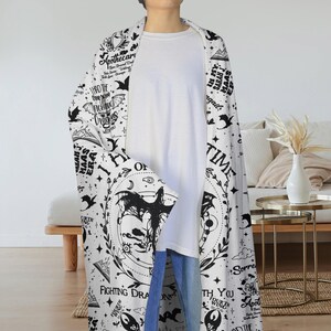 May include: A white and black hooded blanket featuring a dragon design and text. The blanket has a repeating pattern of dragons, stars, and text. The text includes phrases like "Fighting Dragons" and "In My Sarah J Maas Era".