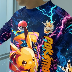 May include: A long-sleeve shirt featuring a vibrant blue background with a graphic of a yellow electric creature, a red and white ball, and bright pink and cyan lightning bolts. The text "THUNDER" and "POKEMON" are printed in yellow.