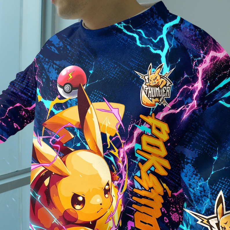 May include: A long-sleeve shirt featuring a vibrant blue background with a graphic of a yellow electric creature, a red and white ball, and bright pink and cyan lightning bolts. The text "THUNDER" and "POKEMON" are printed in yellow.