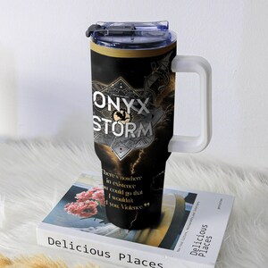 May include: A black and gold travel mug with the text "ONYX STORM" and a quote. The mug has a white handle and a clear lid. The design includes a lightning bolt and a dragon. The mug is sitting on a book.