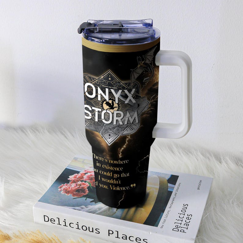 May include: A black and gold travel mug with the text "ONYX STORM" and a quote. The mug has a white handle and a clear lid. The design includes a lightning bolt and a dragon. The mug is sitting on a book.