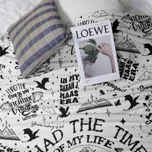 May include: A white blanket with black dragon and text designs, including "I had the time of my life fighting dragons with you." A blue and grey plaid pillow and a "LOEWE" magazine are on top of the blanket.
