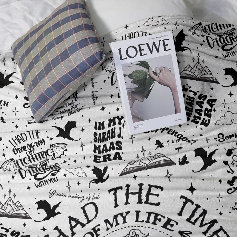 May include: A white blanket with black dragon and text designs, including "I had the time of my life fighting dragons with you." A blue and grey plaid pillow and a "LOEWE" magazine are on top of the blanket.