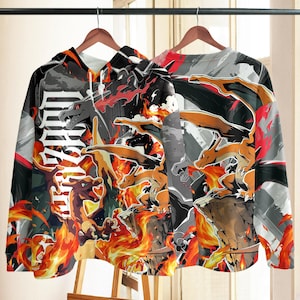 May include: A long-sleeve hooded sweatshirt featuring an all-over graphic print of the orange dragon-like creature Charizard. The design includes flames, rocky terrain, and the text Charizard in a bold white font against a black and gray background.