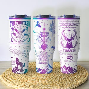 Puede incluir: Tres vasos aislados blancos con tapas negras, cada uno con un diseño único en morado, turquesa y azul. Los diseños incluyen texto, una brújula, un lobo, un ciervo e ilustraciones de fantasía. Los vasos están sobre una estera tejida.