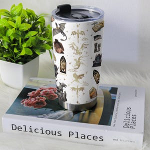 May include: A white stainless steel tumbler with a black lid, featuring a repeating pattern of dragons, books, and gothic window illustrations in gold and black. The tumbler is cylindrical and sits on a book titled "Delicious Places".
