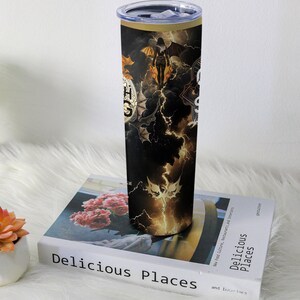 May include: A black and gold stainless steel tumbler with a lid. The tumbler features a design with a woman with wings, bats, lightning, and the text "HGS". The tumbler is sitting on a book titled "Delicious Places".