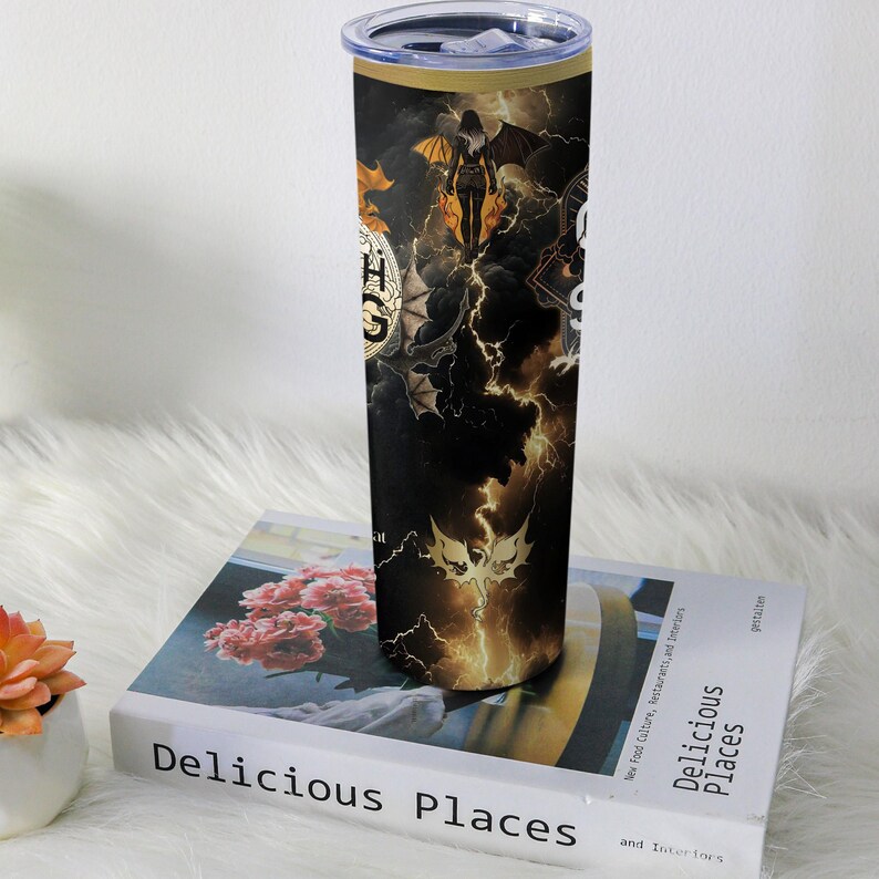 May include: A black and gold stainless steel tumbler with a lid. The tumbler features a design with a woman with wings, bats, lightning, and the text "HGS". The tumbler is sitting on a book titled "Delicious Places".