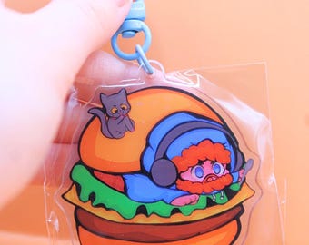 May include: A clear acrylic keychain featuring a cartoon illustration of a person in a blue outfit and headphones, lying on a burger. A gray cat sits on top of the burger. The keychain has a blue clasp.