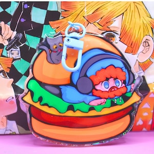Könnte beinhalten: Ein klarer Acryl-Schlüsselanhänger mit einem Cartoon-Charakter in einem Burger. Der Burger hat orange, grüne und rote Schichten. Eine graue Katze sitzt oben, und ein blauer Verschluss ist angebracht. Der Hintergrund zeigt Anime-Kunst.
