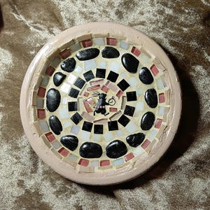 May include: A circular mosaic dish with a pale pink border. The mosaic pattern incorporates black, white, and pink tiles, and a central image of a rabbit with a pocket watch. The dish is likely handmade.