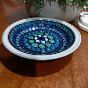 May include: A circular dish with a mosaic tile design. The mosaic features shades of blue and teal tiles, arranged in a circular pattern. The dish has a white rim and is likely ceramic.