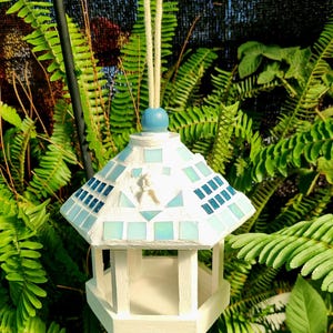 May include: A white, hexagonal bird feeder with a mosaic tile roof in shades of blue and white. It hangs from a rope and blue bead. The background is filled with green foliage.