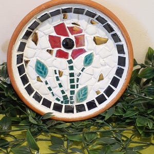 May include: A round terracotta plate featuring a mosaic design of a red flower with a black centre, green leaves, and a stem. The background is composed of white and gold-toned glass pieces, with a border of black square tiles around the edge.