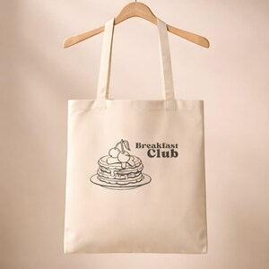 Könnte beinhalten: Cremefarbene Tragetasche an einem Holzbügel. Die Tasche zeigt eine schwarze Illustration von Pfannkuchen mit Kirschen und den Worten "Breakfast Club."