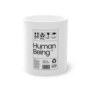 Könnte beinhalten: Weißer Keramikbecher mit dem schwarzen Schriftzug "Human Being", zusammen mit Versandzeichen und dem Text "100% Organic". Der Becher hat eine zylindrische Form und ein schlichtes Design.