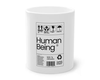 Può includere: Tazza in ceramica bianca con la scritta "Human Being" in nero, insieme a simboli di spedizione e al testo "100% Organic". La tazza ha una forma cilindrica e un design semplice.