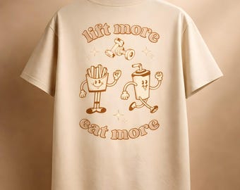 Puede incluir: Camiseta beige con un gráfico marrón de una pesa, patatas fritas y una bebida, con las palabras "lift more" y "eat more". La camiseta está colgada en una percha de madera.