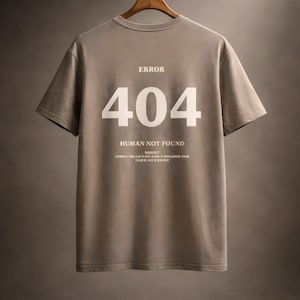 Könnte beinhalten: Beigefarbenes T-Shirt mit dem weißen Aufdruck „ERROR 404 HUMAN NOT FOUND“. Darunter steht der Satz „SORRY: SURE THAN YOU ARE LOOKING FOR DOES NOT EXIST“. Das T-Shirt hängt an einem Holzkleiderbügel vor neutralem Hintergrund.