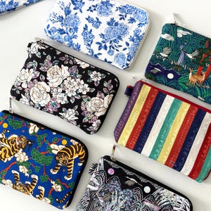 May include: A collection of zippered pouches with various patterns. Designs include blue floral, black and white roses, a tiger scene, a landscape with deer, and a striped pattern. Each pouch has a different color scheme and design.