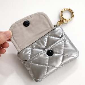 May include: Silver quilted coin purse with a gold-colored keychain. The purse has a diamond pattern and a snap closure. The interior is a light beige color. The purse is open, revealing the interior.