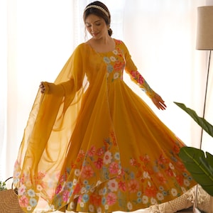 May include: Mustard yellow Anarkali suit with floral print. The long-sleeved dress features a round neckline and a flowing skirt. The floral pattern includes pink, blue, and white flowers. A matching sheer dupatta and pants complete the ensemble.