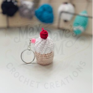 May include: A crocheted cupcake keychain with a red cherry on top. The cupcake has a white top and a tan base. A pink pom-pom and a silver keyring are attached. Other crocheted items are blurred in the background.