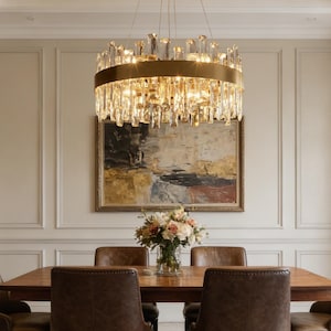 May include: A luxurious chandelier with a gold-toned circular frame and numerous crystal accents hangs above a wooden dining table. The crystals are arranged in vertical rows, creating a sparkling effect. The room features a painting and a floral arrangement.