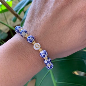 May include: A white bracelet with blue floral designs and gold accents. The bracelet has a heart charm with a gold outline.