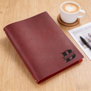 May include: A deep red leather notebook cover featuring a black engraved letter B with the name Braxton centered across the middle. The cover has a smooth matte texture and is positioned next to a white coffee mug and a black pen on paper.
