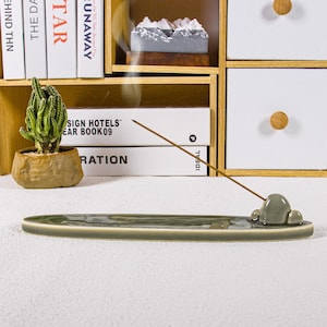 May include: A sage green ceramic incense holder with a cloud-shaped design. The holder is long and oval, with a small, raised cloud detail at one end. An incense stick is lit, emitting a thin stream of smoke. A small cactus in a stone-like pot is visible.