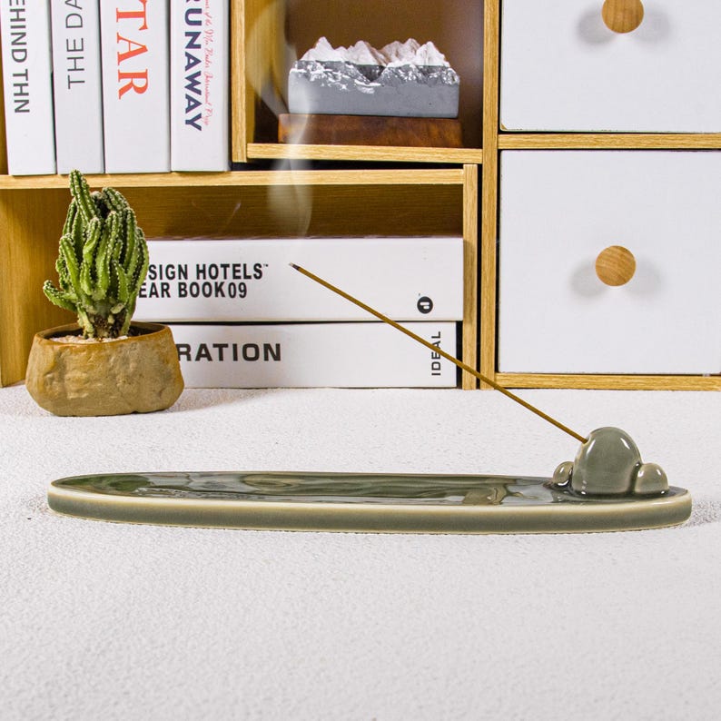 May include: A sage green ceramic incense holder with a cloud-shaped design. The holder is long and oval, with a small, raised cloud detail at one end. An incense stick is lit, emitting a thin stream of smoke. A small cactus in a stone-like pot is visible.