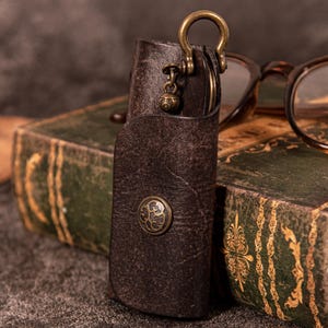 Könnte beinhalten: Ein brauner Leder-Schlüsselhalter mit Messingverschluss und dekorativem Knopf. Der Schlüsselhalter ist offen und zeigt Schlüssel im Inneren. Ein kleiner Messinganhänger hängt am Verschluss. Der Schlüsselhalter liegt auf einem alten Buch.