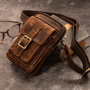 May include: A brown leather crossbody bag with a distressed finish, featuring a front flap secured by a brass buckle. The bag has a zippered main compartment and a brown fabric strap. A pair of eyeglasses and a vintage book are in the background.