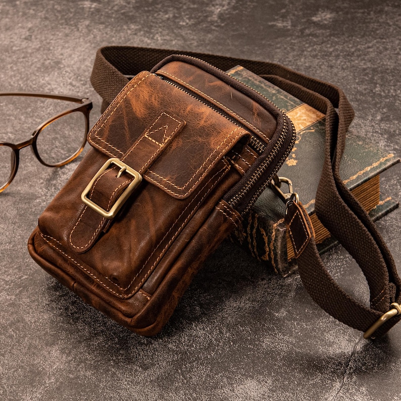 May include: A brown leather crossbody bag with a distressed finish, featuring a front flap secured by a brass buckle. The bag has a zippered main compartment and a brown fabric strap. A pair of eyeglasses and a vintage book are in the background.