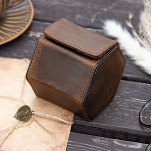 May include: A brown leather watch case with a hexagonal shape and a hinged lid. The case is made of distressed leather with visible stitching and a dark brown trim. The case is sitting on a dark wooden surface.