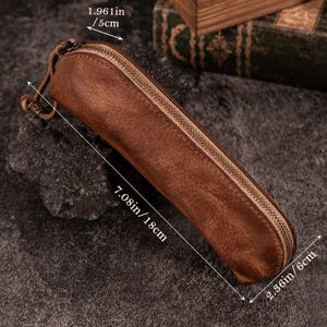 May include: A brown leather pencil case with a zipper closure. The case measures 7.08 inches long, 2.36 inches wide, and 1.96 inches high. The pencil case has a loop on one end.