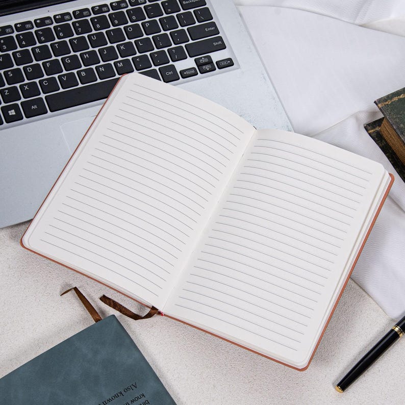 May include: An open, lined notebook with a brown cover and a brown ribbon bookmark. A laptop keyboard is visible in the background. A black pen and a teal notebook are also in the image.