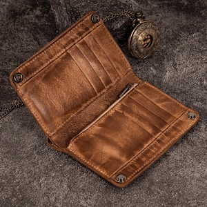 May include: Open brown leather wallet with multiple card slots and a pen holder. The wallet has a distressed finish and is secured with metal snaps. A vintage pocket watch with a chain is in the background.