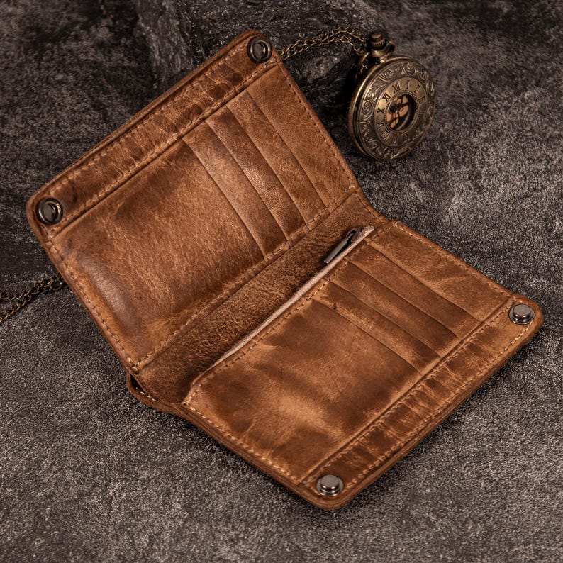 May include: Open brown leather wallet with multiple card slots and a pen holder. The wallet has a distressed finish and is secured with metal snaps. A vintage pocket watch with a chain is in the background.