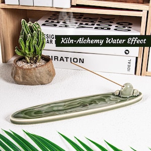 May include: A green ceramic incense holder with a water effect design. The holder has a small, rounded incense stick holder. A small cactus in a brown pot sits to the left. The text "Kiln-Alchemy Water Effect" is visible.