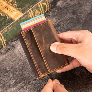 May include: A brown leather wallet with a worn, textured appearance. The wallet is open, revealing a stack of colorful credit cards. A black elastic band secures the wallet's contents. The wallet is held in a person's hand.