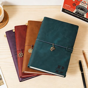 May include: A selection of leather-bound journals in purple, red, brown, and teal. Each journal has a decorative charm and a closure strap. The teal journal has the name "Braxton" engraved on the cover.